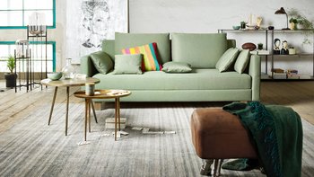 JOKA Comoda Doppelliege in Hellgrau mit bunten Zierkissen in einem modernen Wohnbereich mit Couchtisch, Blumen und dekorativen Kunstwerken an der Wand.