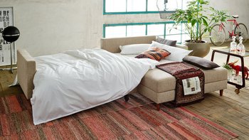 Zur Schlafstätte umgebautes Ecksofa mit weißem Bettzeug und Kissen in moderner Loft-Umgebung.