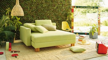 JOKA Quadro Schlafsofa in Hellgrün in geschlossener Position mit Zierkissen in einem Gartenambiente.