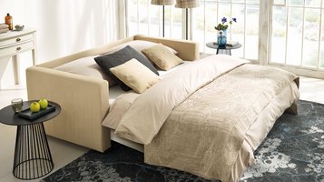 JOKA Magic Längsschläfer umgebaut als komfortables Bett mit beiger Bettwäsche, gemustertem Bezug und dekorativen Kissen in einem hellen Raum mit Teppich und Beistelltisch.