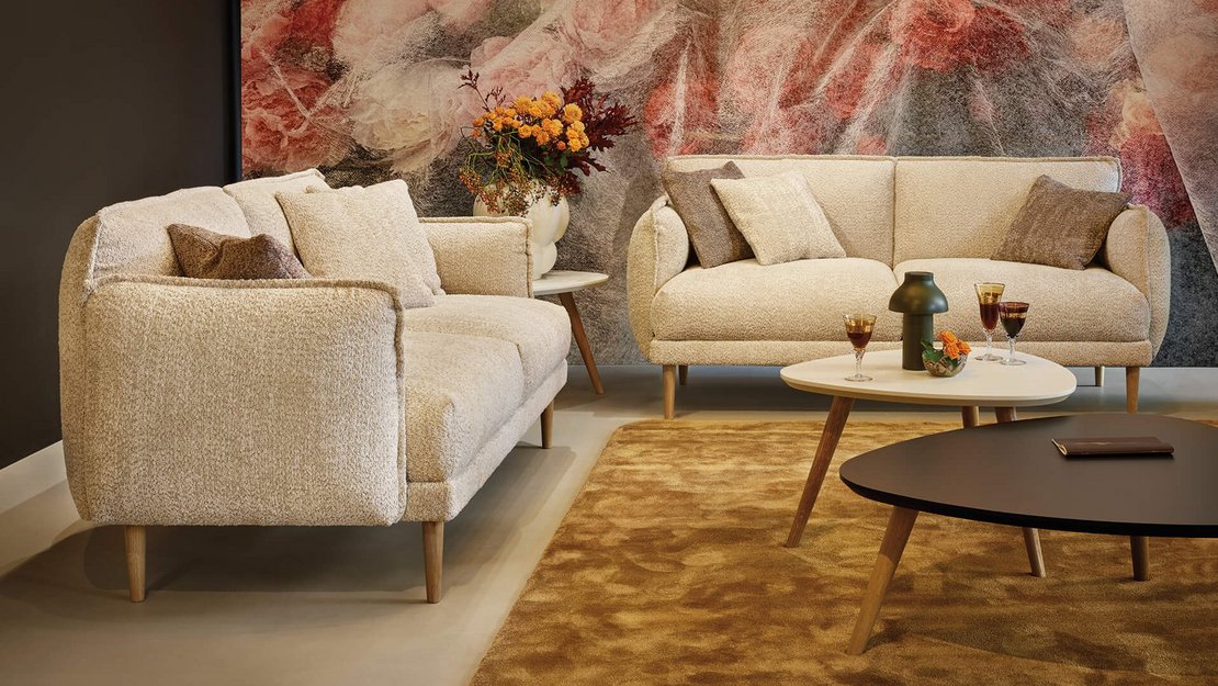 Frontalansicht zweier beigefarbener JOKA Bouclé-Sofas im Loungebereich mit abgestimmten Zierkissen und modernem Beistelltisch-Ensemble.
