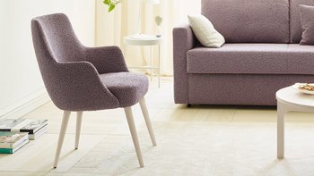 JOKA Paolo Stuhl mit violettem Stoffbezug und weiß lasierten Holzfüßen neben einem passenden Sofa in einem hellen Wohnzimmer.
