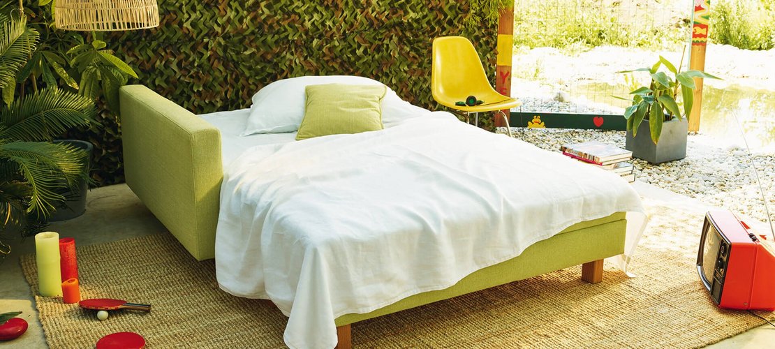 JOKA Quadro Schlafsofa in Hellgrün, ausgeklappt als Bett mit weißer Bettwäsche und Kissen.