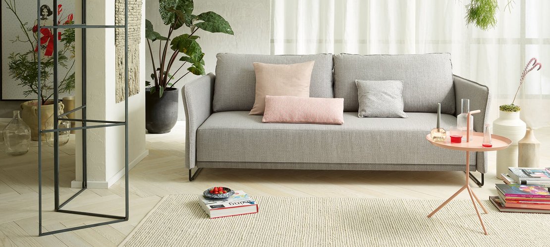 Hellgraue JOKA Lino Doppelliege mit passenden rosa und grauen Zierkissen in einem modernen Wohnbereich mit hellem Teppich und Beistelltisch.