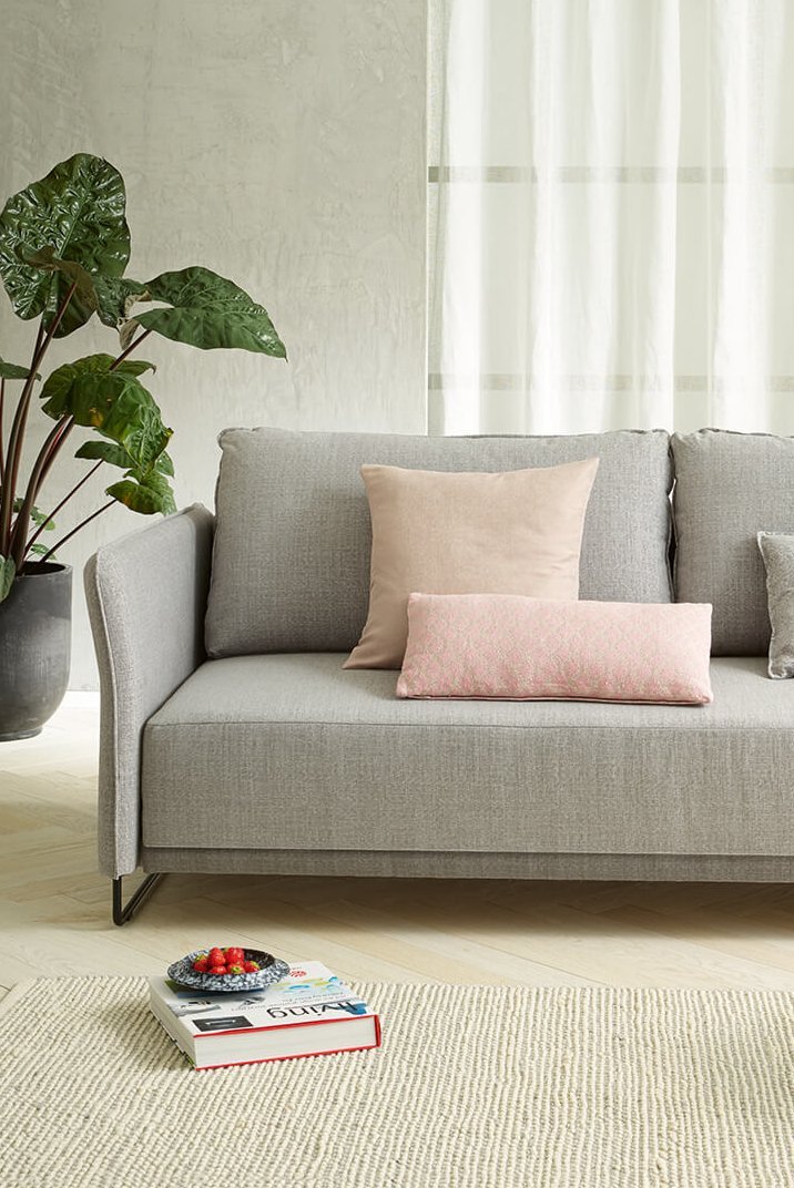 Hellgraue JOKA Lino Doppelliege mit passenden rosa und grauen Zierkissen in einem modernen Wohnbereich mit hellem Teppich und Beistelltisch.