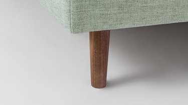 Nahaufnahme eines geraden Kegelfusses aus Holz an einem hellgrünen Sofa-Unterbau