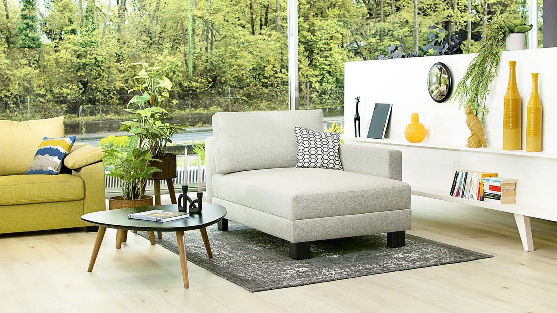 Hellgraue JOKA Quadro Relaxliege in einem lichtdurchfluteten Wohnzimmer mit großen Fensterfronten und moderner Dekoration.