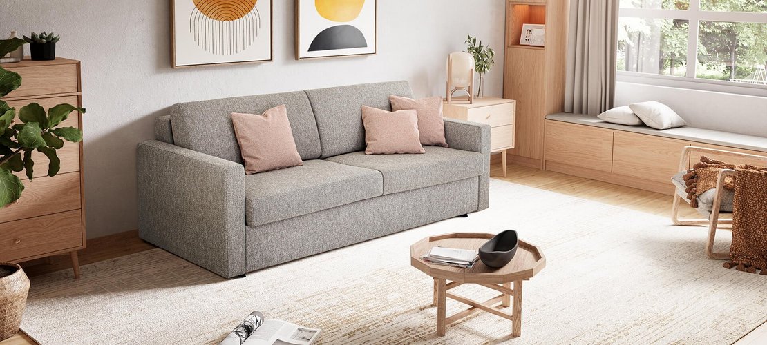 JOKA Marea Längsschläfer in Grau mit drei rosafarbenen Zierkissen in einem hellen, modern eingerichteten Wohnraum mit Teppich und großen Fenstern.