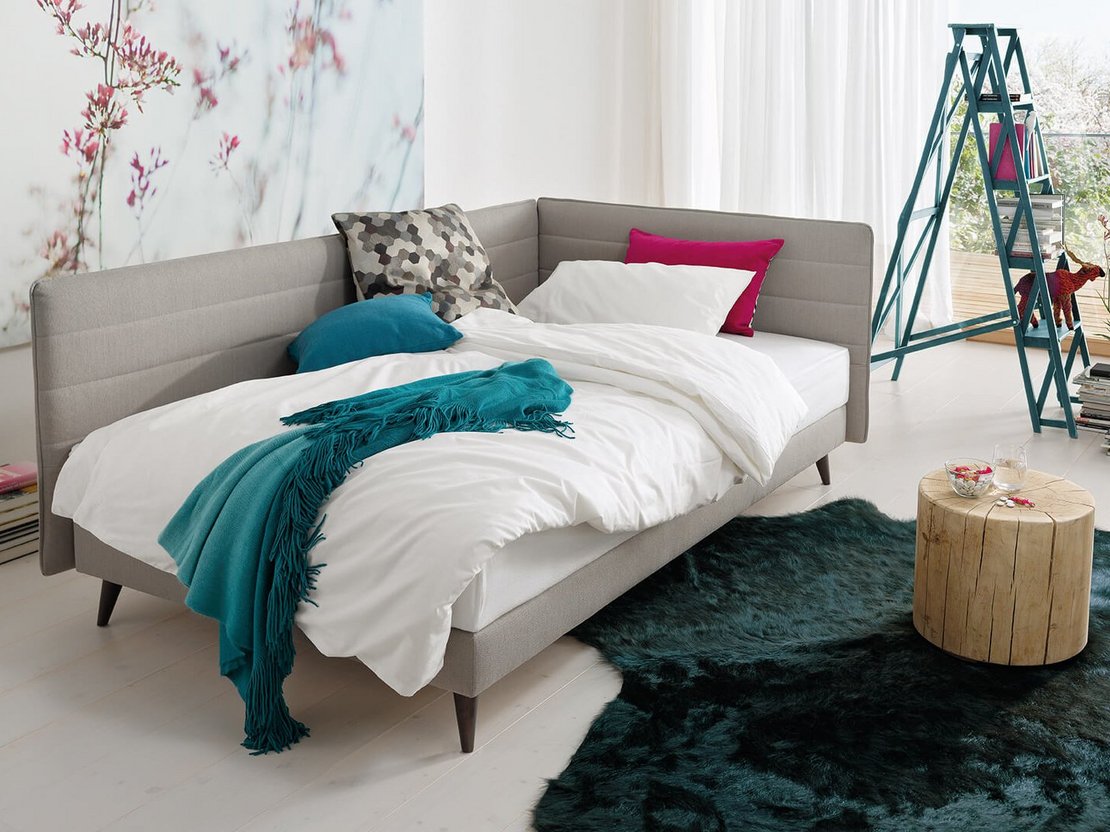 Als Bett hergerichtete grau-beige JOKA Toscana Einzelliege mit weißem Bettzeug und bunten Kissen in einem hellen Wohnraum