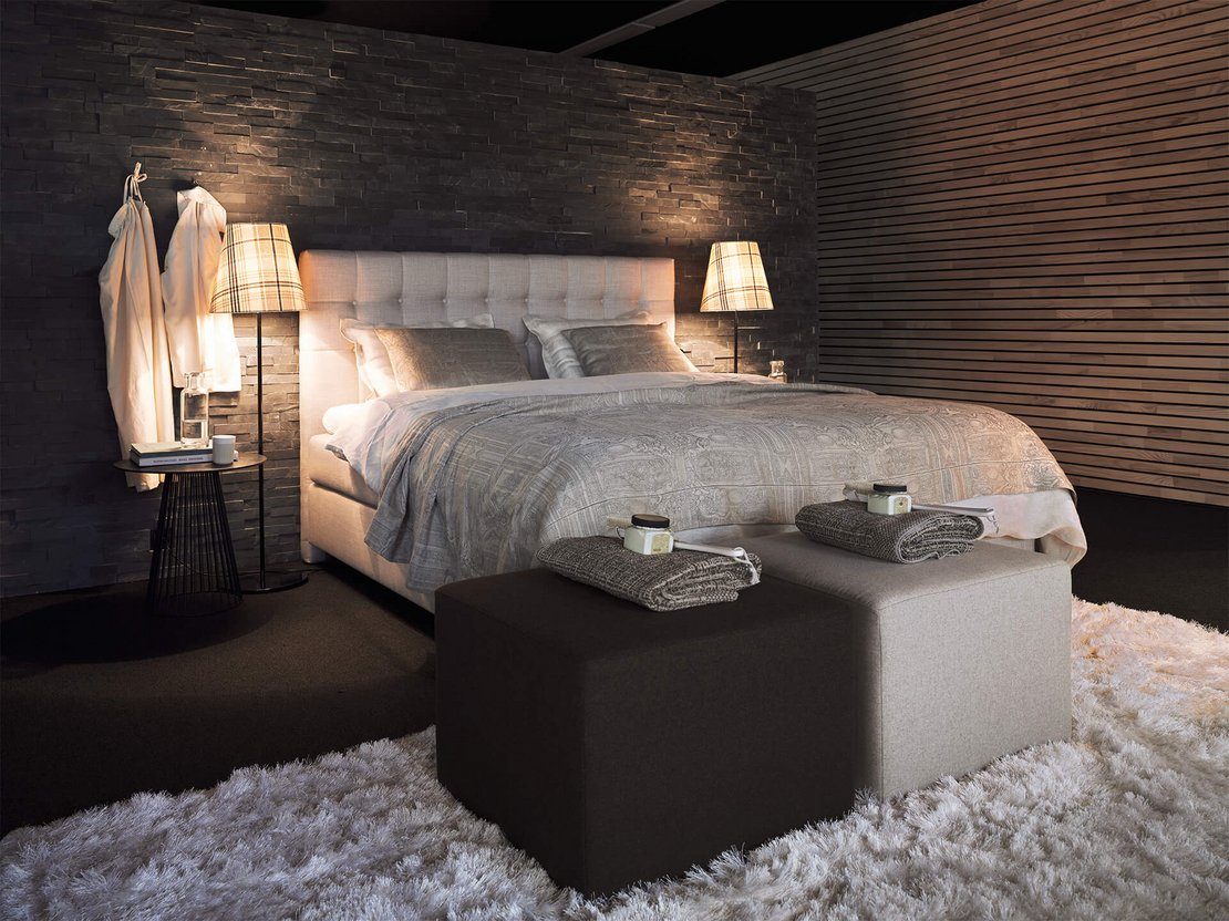 Hellgraues JOKA Noblesse Boxspringbett in dunklem Wellness-Ambiente mit Holz- und Steinwand