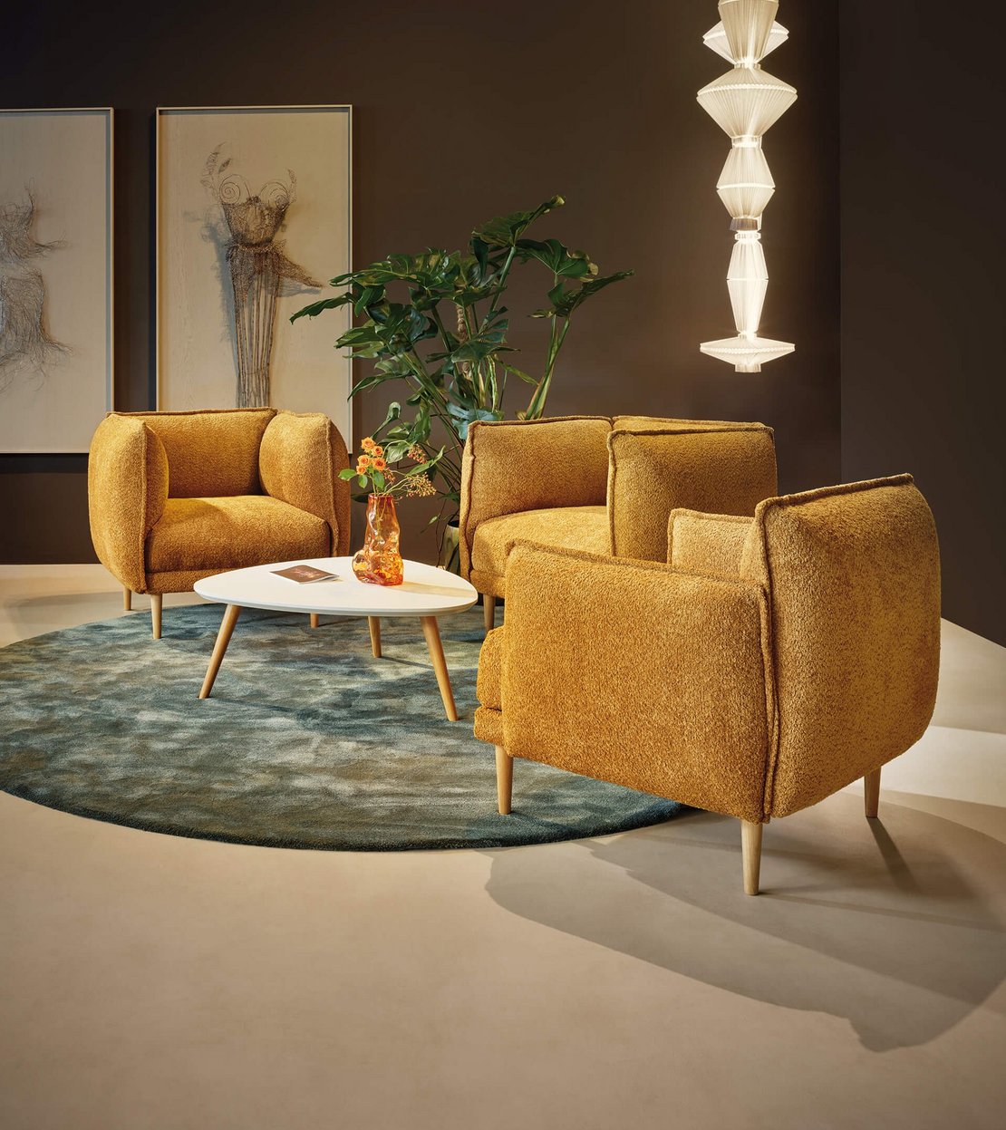 Sitzgruppe aus drei gelben JOKA Bouclé-Sesseln um einen hellen Beistelltisch auf rundem grünem Teppich neben einer Zimmerpflanze und modernem Lichtdesign.