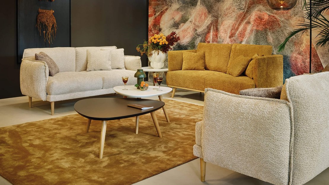 JOKA Loungebereich mit beigefarbenem und senfgelbem Sofa, Bouclé-Sessel und zwei nierenförmigen Beistelltischen auf braunem Teppich vor einer Wand mit Blumenmotiv.