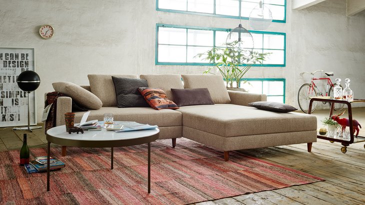 Modernes Loft-Wohnzimmer mit einer beigen Eckcouch inklusive Recamiere, rundem Couchtisch und verputzter Wand im Hintergrund.