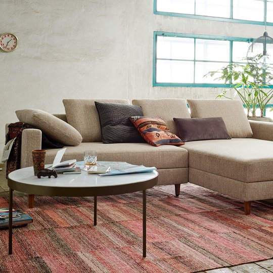 Modernes Loft-Wohnzimmer mit einer beigen Eckcouch inklusive Recamiere, rundem Couchtisch und verputzter Wand im Hintergrund.