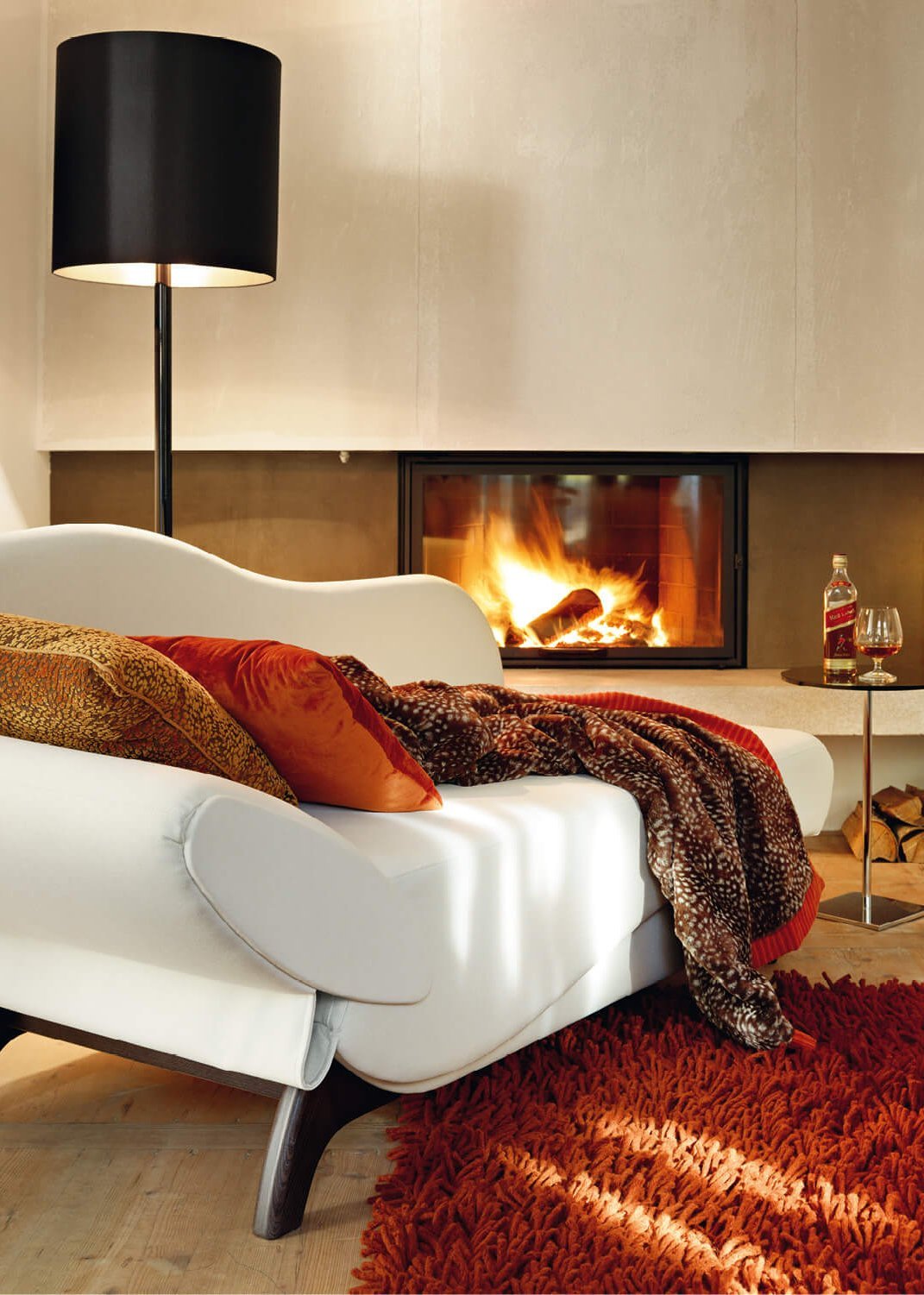 Weiße JOKA Classique Chaiselongue mit roten Kissen und Decke in einem gemuetlich eingerichteten Zimmer vor einem brennenden Kamin.