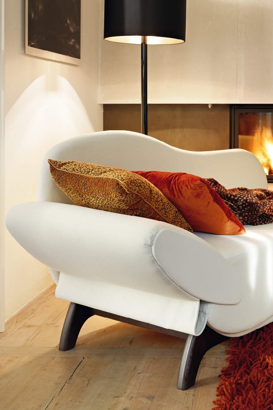 Weiße JOKA Classique Chaiselongue mit roten Kissen und Decke in einem gemuetlich eingerichteten Zimmer vor einem brennenden Kamin.