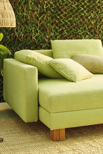 JOKA Quadro Schlafsofa in Hellgrün in geschlossener Position mit Zierkissen in einem Gartenambiente.