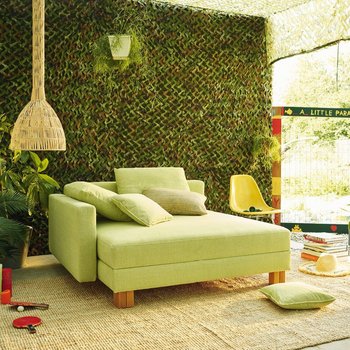 JOKA Quadro Schlafsofa in Hellgrün in geschlossener Position mit Zierkissen in einem Gartenambiente.