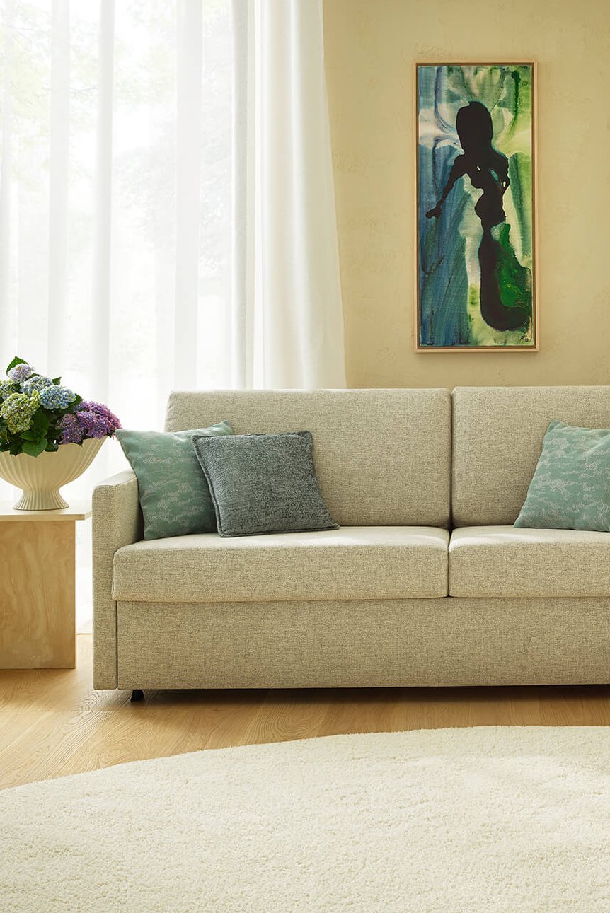 Ansicht eines eleganten Hotelzimmers mit einem beigefarbenen JOKA Sofa mit türkisblauen Akzentkissen vor großen Fenstern und dem Fußende eines passenden türkisblauen Betts.