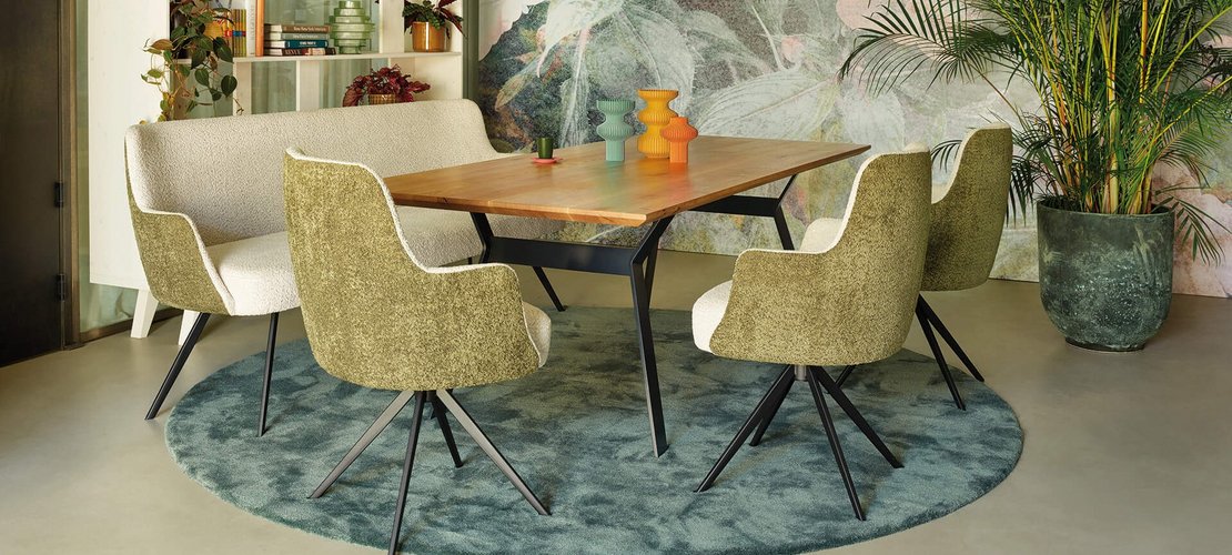 Elegantes JOKA Speiseseparee mit Holztisch, dreifarbigen Vasen, zweifarbiger Polsterbank und Armlehnenstühlen auf rundem blau-grünem Teppich vor einer floralen Fototapete.