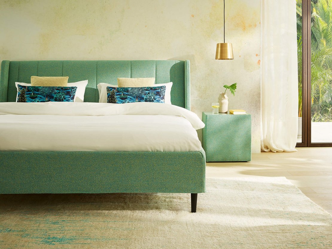 Gepolstertes JOKA Fiore Bett in Salbeigrün mit passendem Nachttisch und goldener Hängeleuchte im Hochformat.