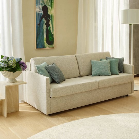 Beigefarbenes JOKA 2-Sitzer Sofa mit grün-blauen Zierkissen auf hellem Holzboden und rundem Teppich neben einer Stehlampe und Blumenbeistelltisch.