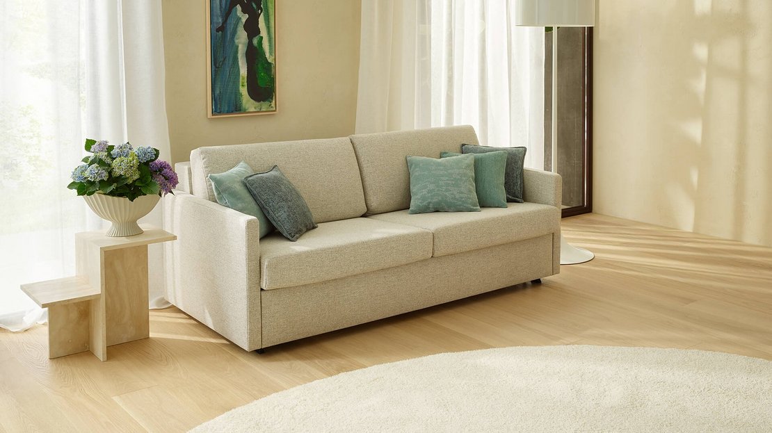 Beigefarbenes JOKA 2-Sitzer Sofa mit grün-blauen Zierkissen auf hellem Holzboden und rundem Teppich neben einer Stehlampe und Blumenbeistelltisch.