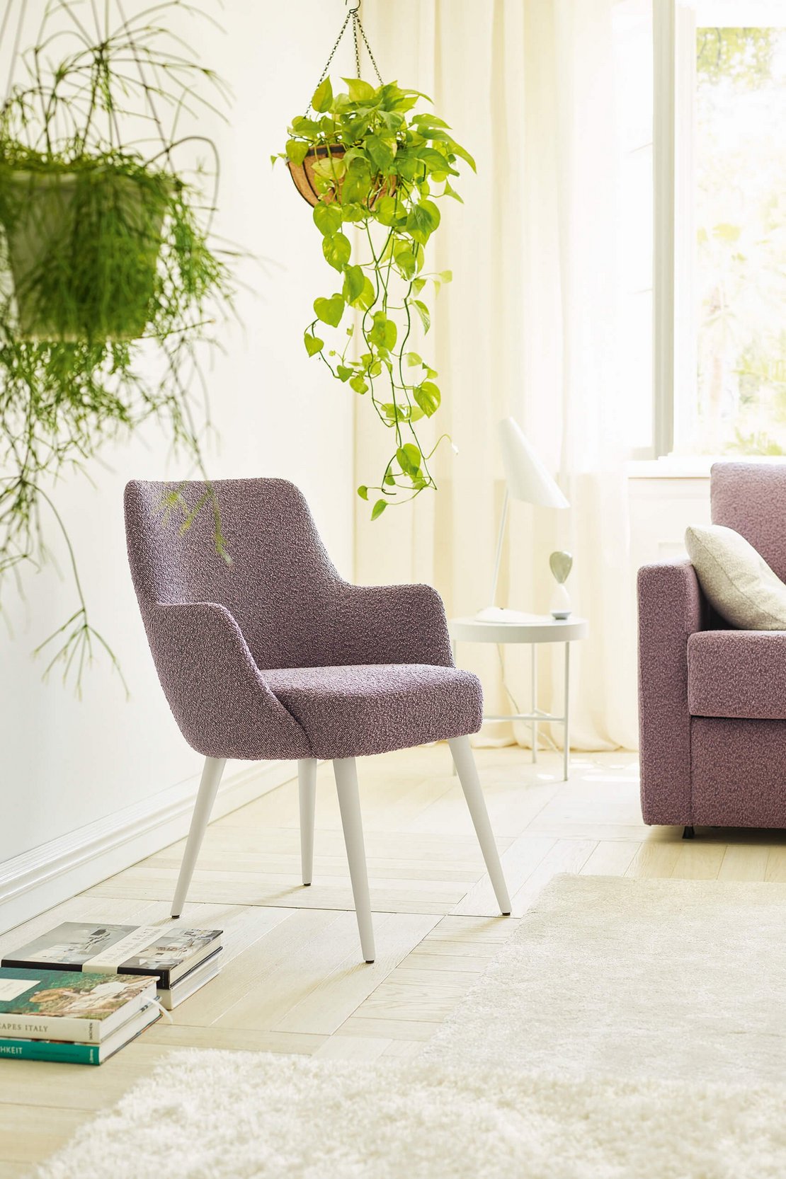 JOKA Paolo Stuhl mit violettem Stoffbezug und weiß lasierten Holzfüßen neben einem passenden Sofa in einem hellen Wohnraum mit Pflanzen.