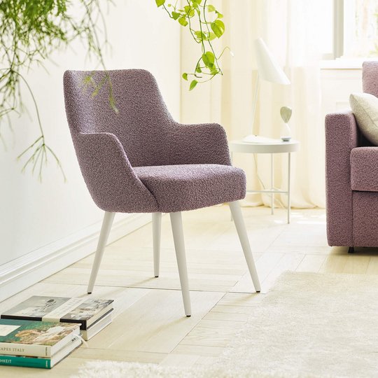 JOKA Paolo Stuhl mit violettem Stoffbezug und weiß lasierten Holzfüßen neben einem passenden Sofa in einem hellen Wohnraum mit Pflanzen.