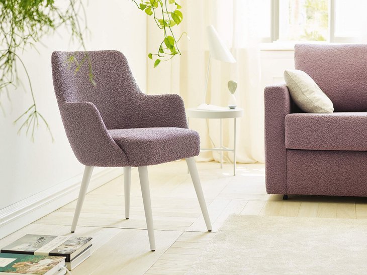 JOKA Paolo Stuhl mit violettem Stoffbezug und weiß lasierten Holzfüßen neben einem passenden Sofa in einem hellen Wohnraum mit Pflanzen.