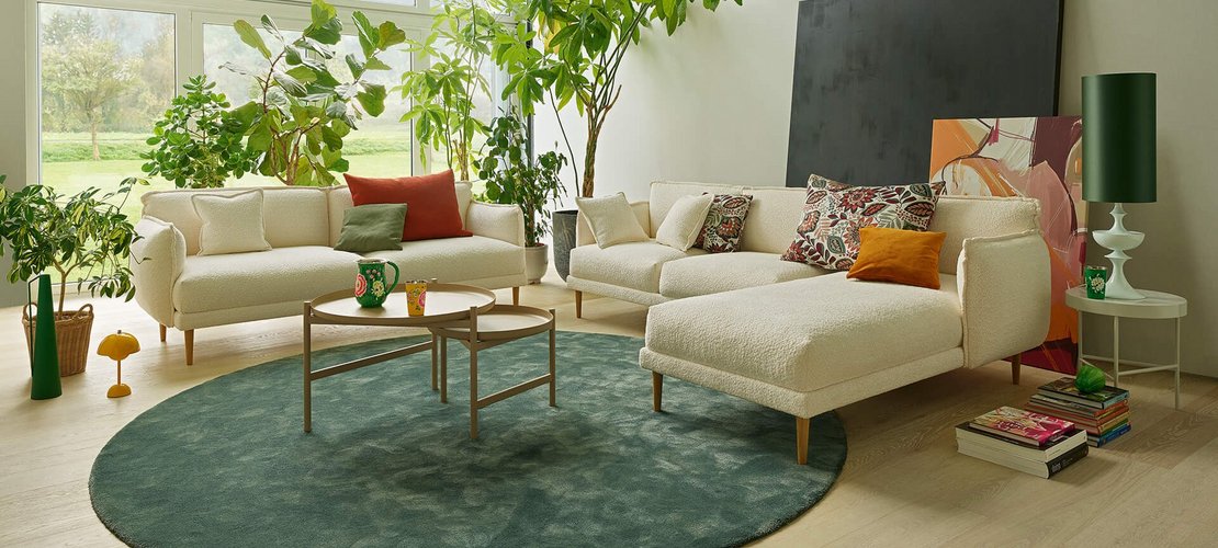 Heller Wohnbereich mit zwei Bouclé-Sofas (3-Sitzer und 2-Sitzer mit Chaiselongue), Couchtisch, grünem Teppich und großen Zimmerpflanzen.