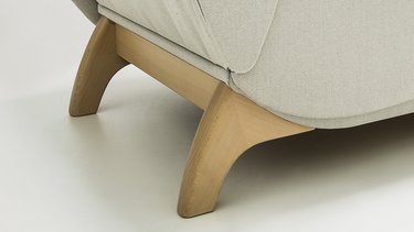 `Nahaufnahme der geschwungenen Holzfüße an der hellgrauen JOKA Classique Chaiselongue vor weißem Hintergrund.`