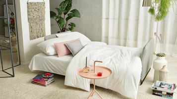 JOKA Lino Doppelliege umgebaut als gemütliches Bett mit weißer Bettwäsche, rosa und grauen Kissen in einem hellen Wohnraum.