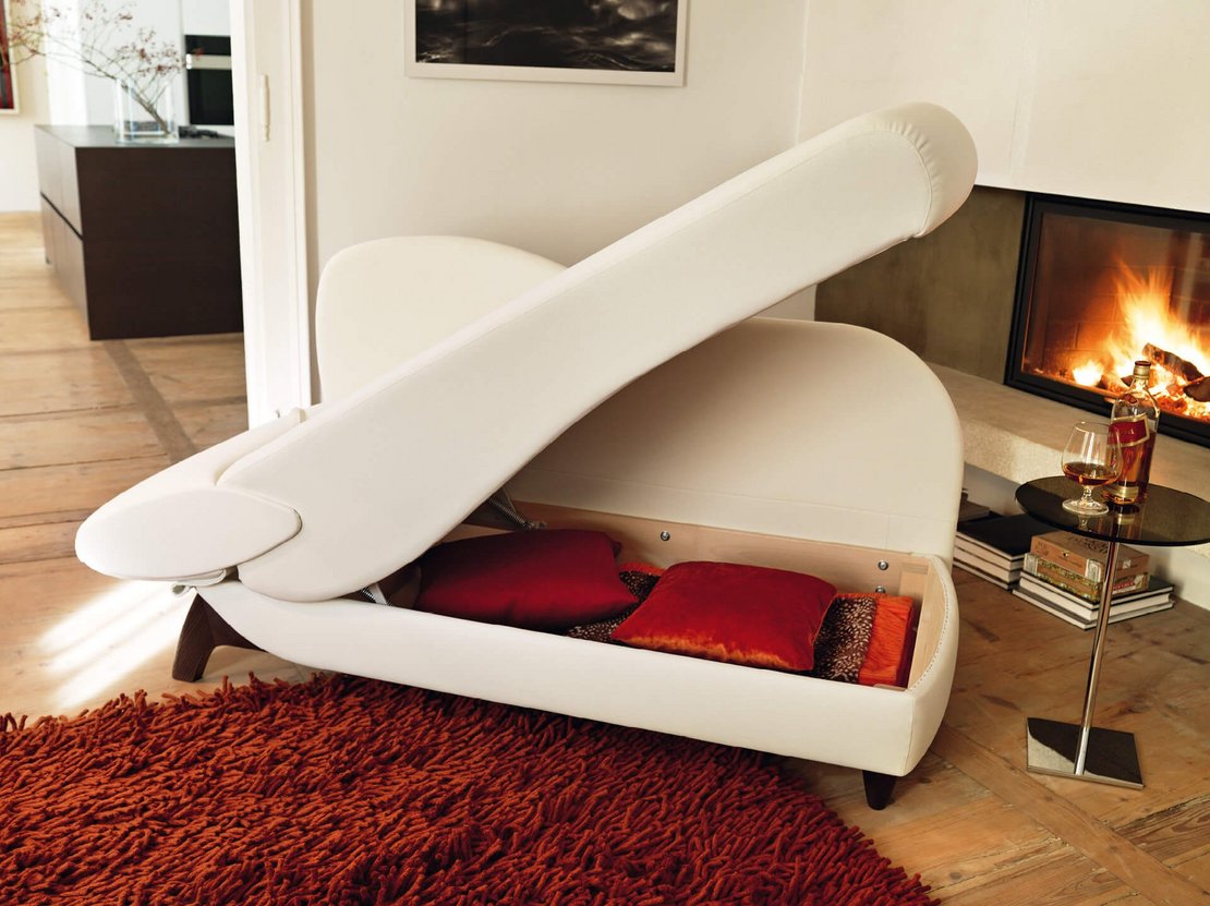Nahaufnahme der hochgeklappten Sitzfläche der weißen JOKA Classique Chaiselongue, um den integrierten Stauraum mit roten Kissen im Inneren zu zeigen.