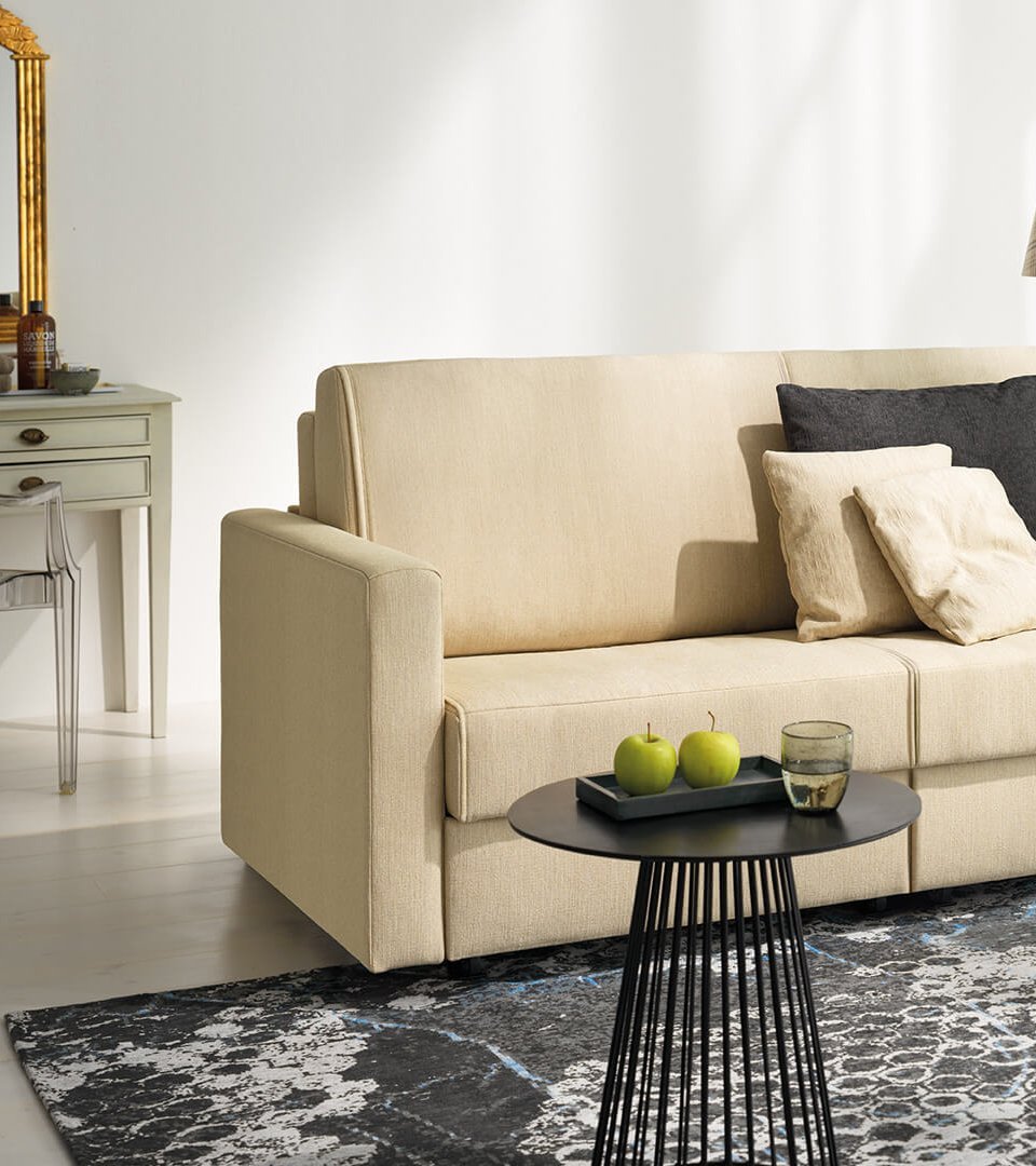 joka-magic-laengsschlaefer.jpg: Beige JOKA Magic Längsschläfer Couch dekoriert mit dunklen und hellen Zierkissen in einem hellen Wohnraum mit gemustertem Teppich und schwarzen Beistelltischen.