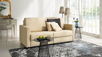 joka-magic-laengsschlaefer.jpg: Beige JOKA Magic Längsschläfer Couch dekoriert mit dunklen und hellen Zierkissen in einem hellen Wohnraum mit gemustertem Teppich und schwarzen Beistelltischen.