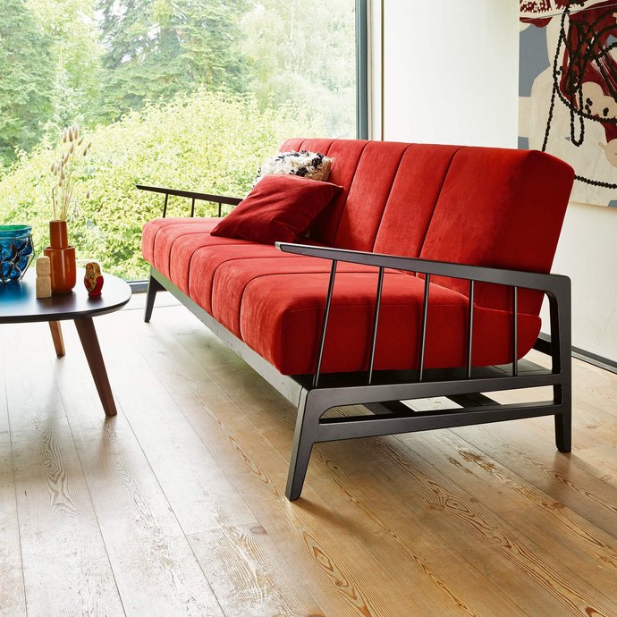 JOKA Cento Kippcouch in Rot mit schwarzem Holzgestell, aufgenommen im Hochformat mit Fokus auf die Armlehne vor einer großen Fensterfront und einem abstrakten Kunstwerk.